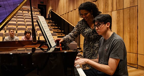 A teacher stands next to a student who is sat at a piano as two other students observe